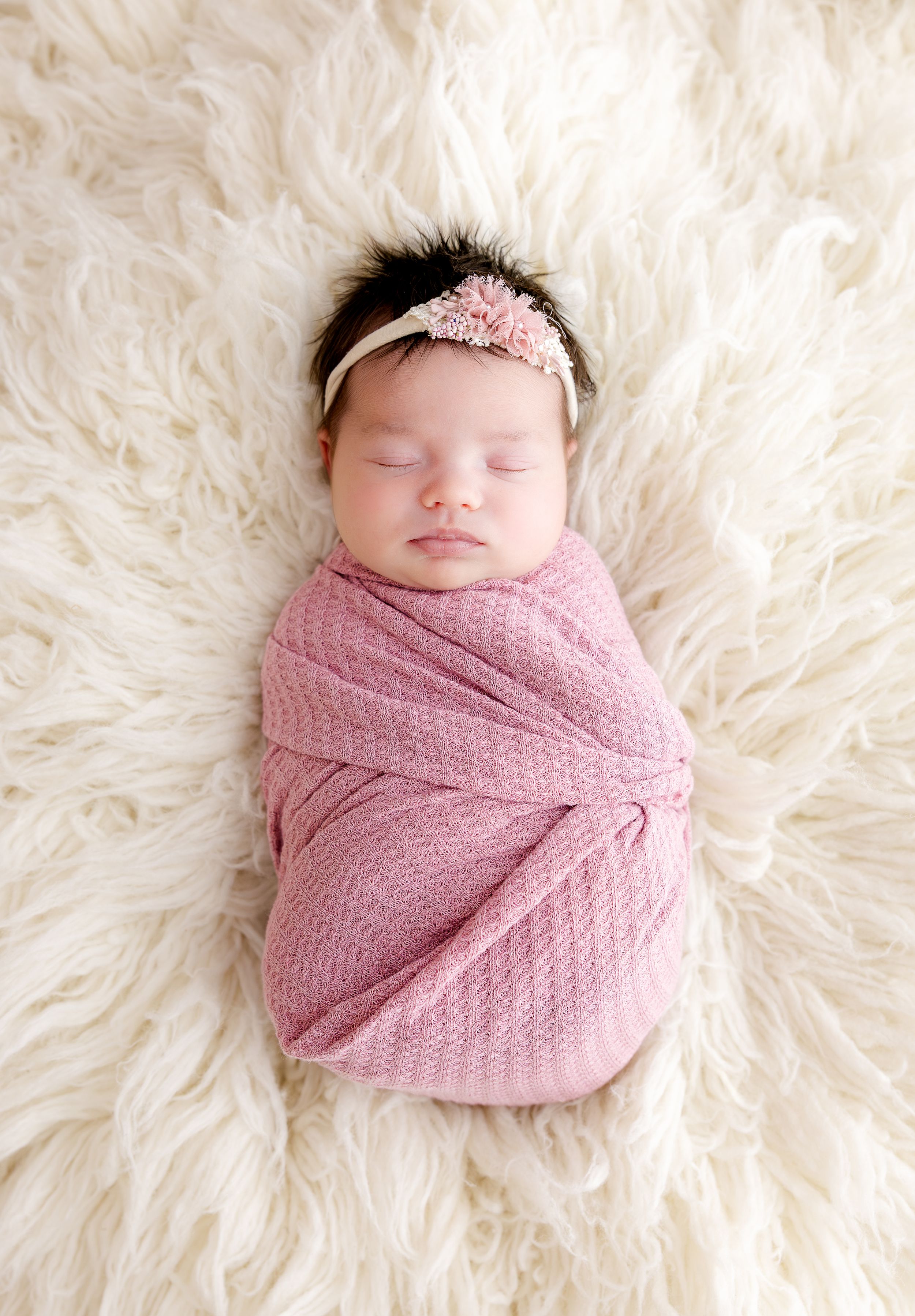 Sleeping newborn wrapped in pink textured swaddle blanket lying on white fluffy rug with matching pink headband.