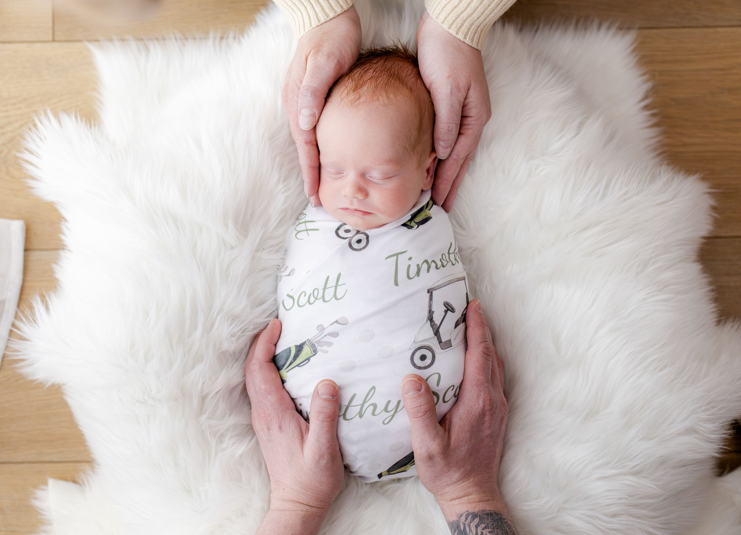 Newborn baby swaddled in a personalized name blanket, cradled by parents hands on a white fluffy rug.
