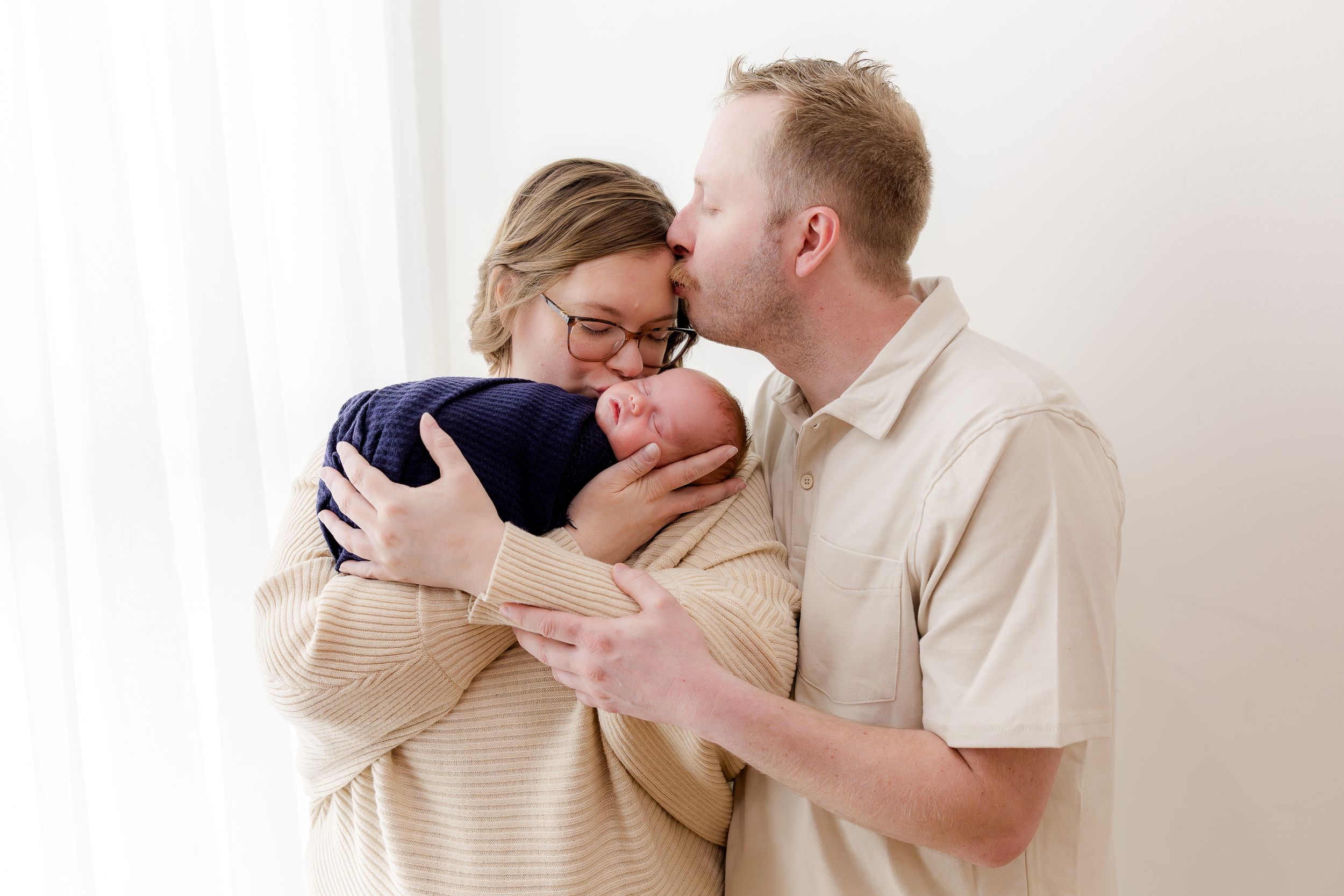 A couple shares a tender moment holding their swaddled newborn baby near a bright window in a studio portrait.