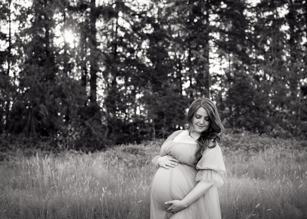 A black and white maternity photo shows an expectant mother in a flowing dress standing in a forest clearing.