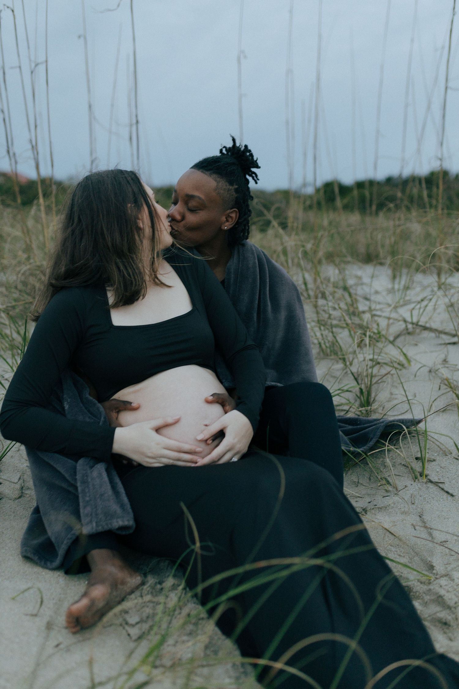 Intimate couple sharing tender moment on beach sand with pregnant belly and dune grass in moody lighting.