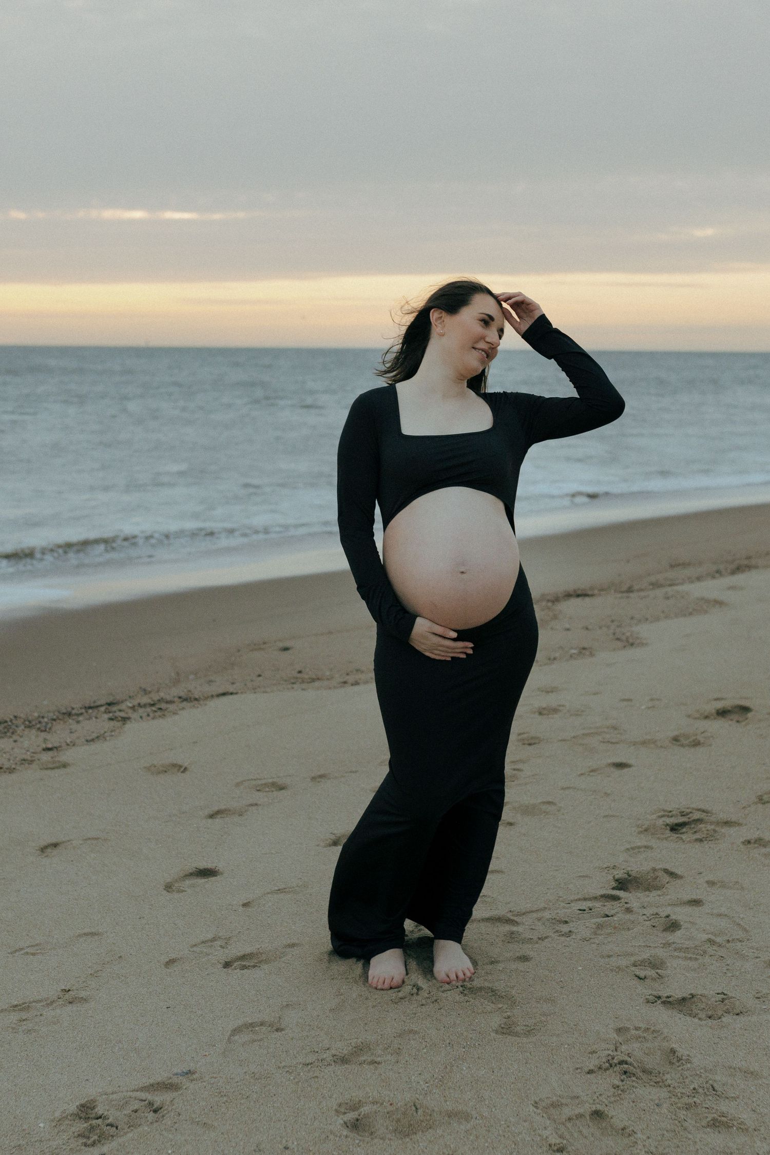 An expectant mother in a black dress poses gracefully on a sunset beach as waves roll in behind her.