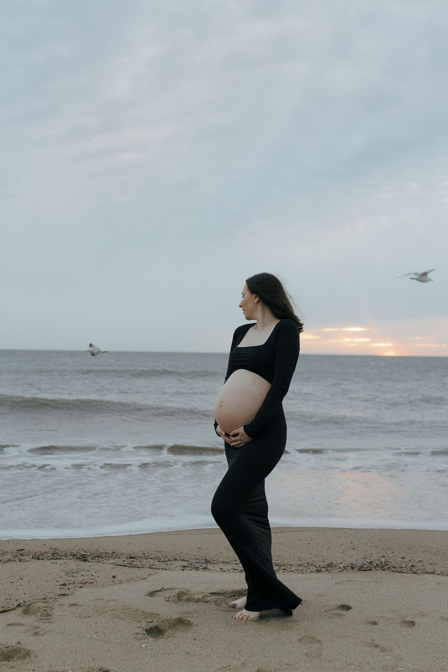 A pregnant woman in black clothing stands on a beach at sunset with seagulls flying in the background.