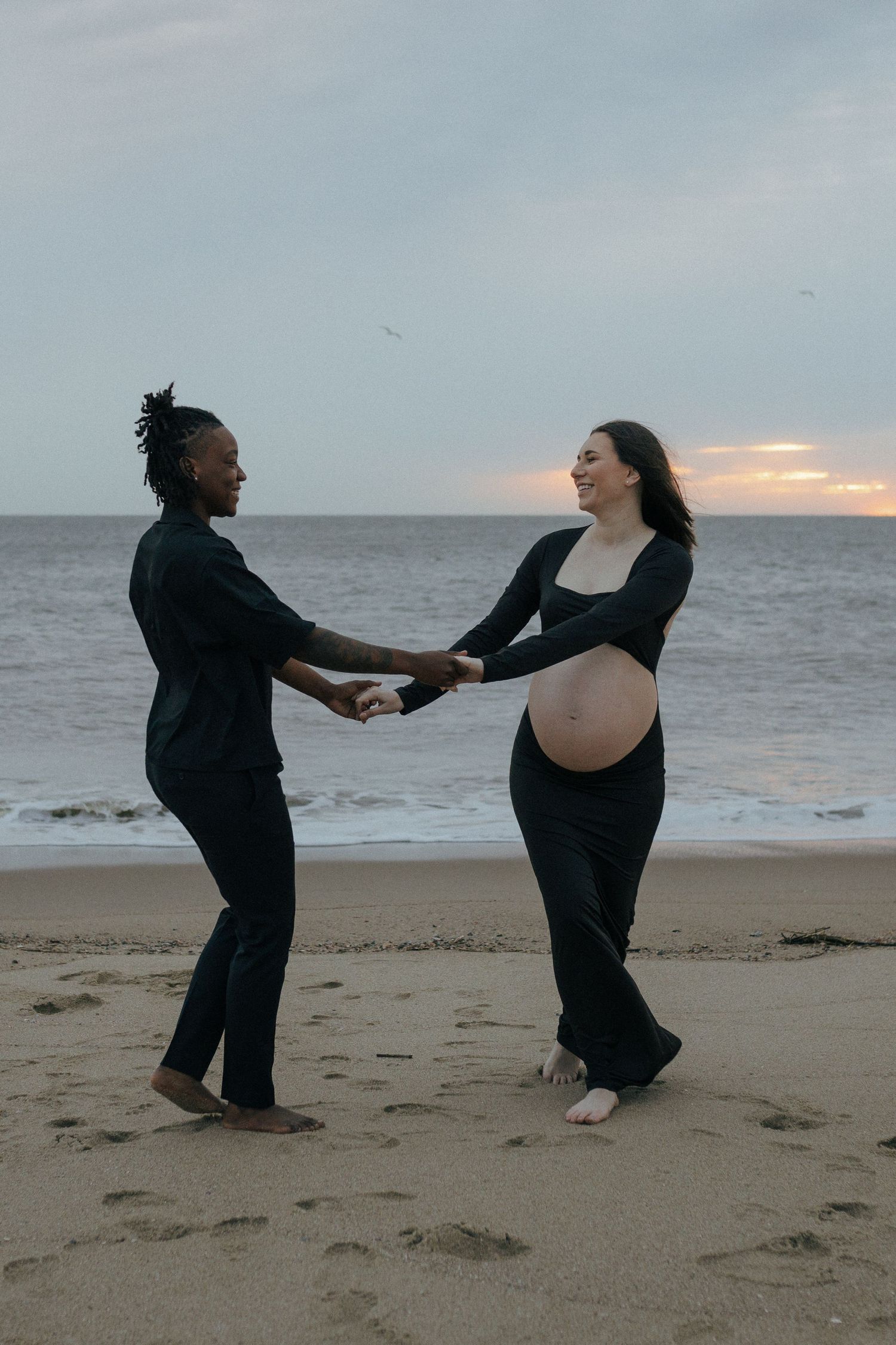 A couple dances together on a beach at sunset as they celebrate their pregnancy with the expectant mother showing her baby bump.