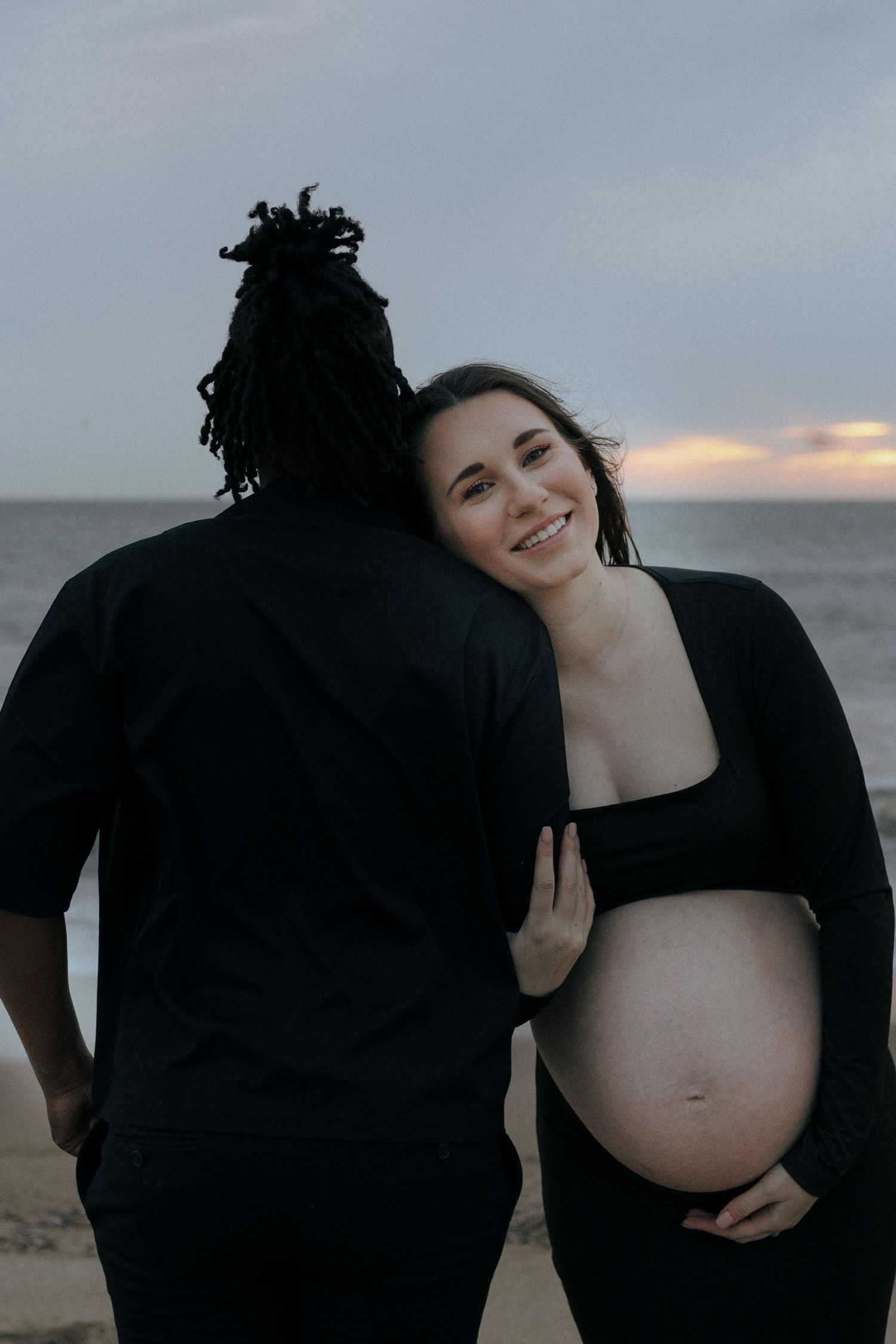 A couple stands together at sunset on the beach, with the pregnant woman wearing black and lovingly holding her belly.