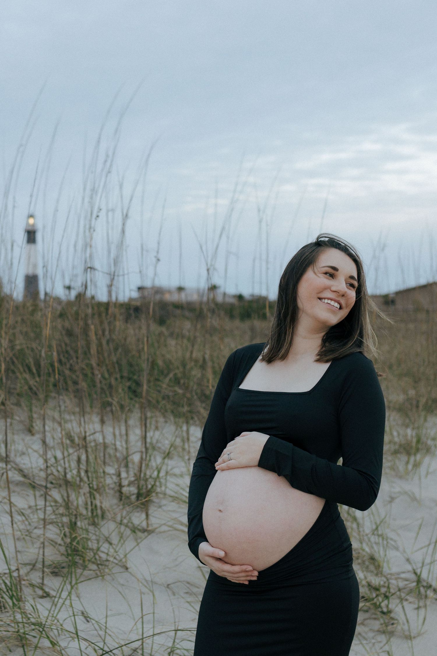 Expecting mother in black dress stands on beach dunes with lighthouse silhouetted in background at dusk.