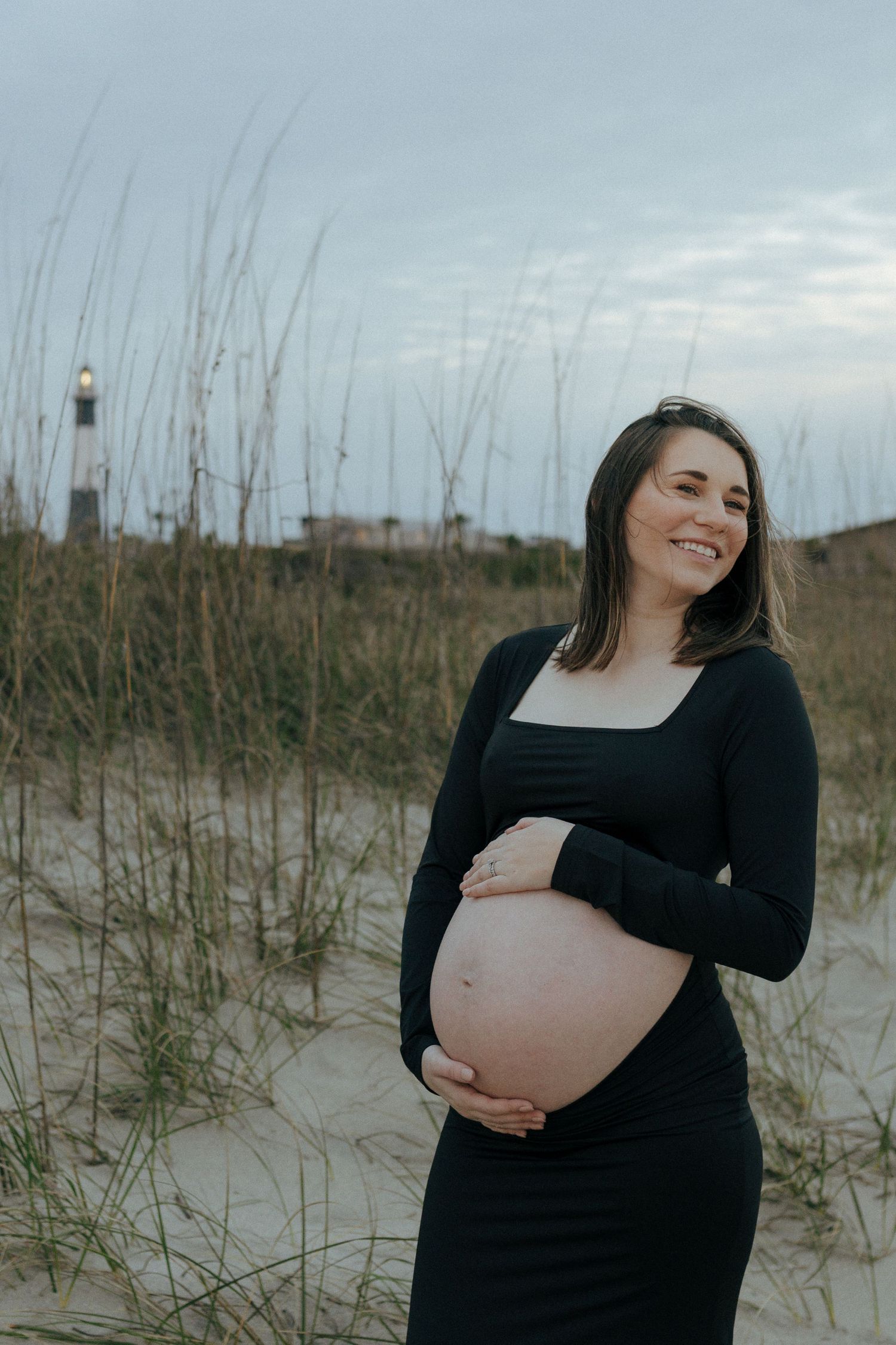 A pregnant woman in a black dress stands on sand dunes with a lighthouse visible in the background at dusk.