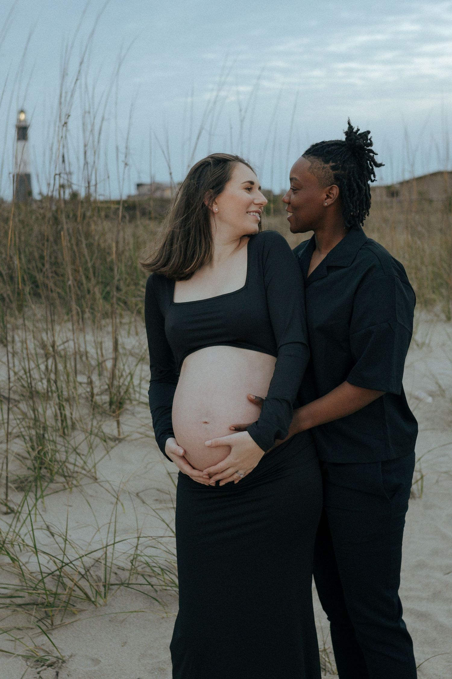 Loving couple in black outfits share intimate maternity moment on sandy beach with lighthouse in background.