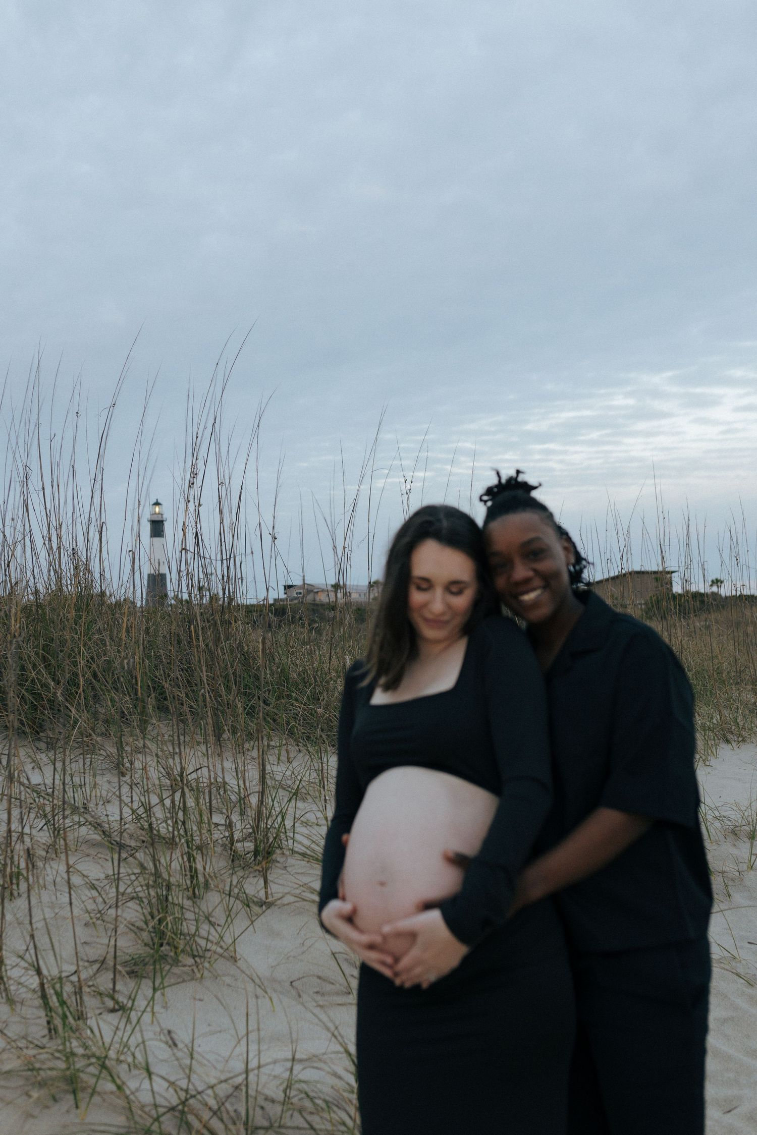 A couple stands together on a sandy beach with tall grass and a lighthouse visible in the background during sunset.