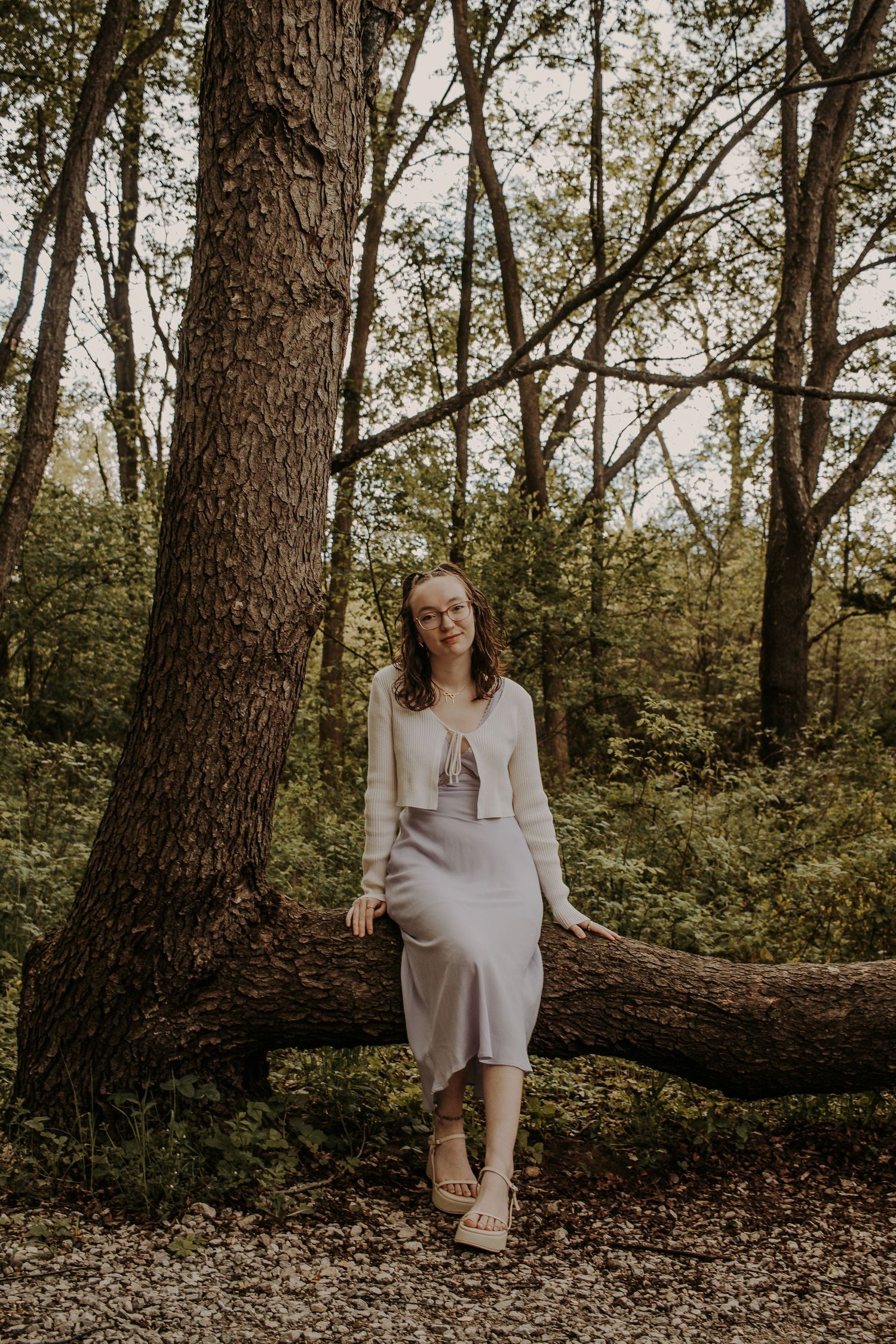 Serene forest photoshoot with person in cream outfit sitting on fallen tree trunk.