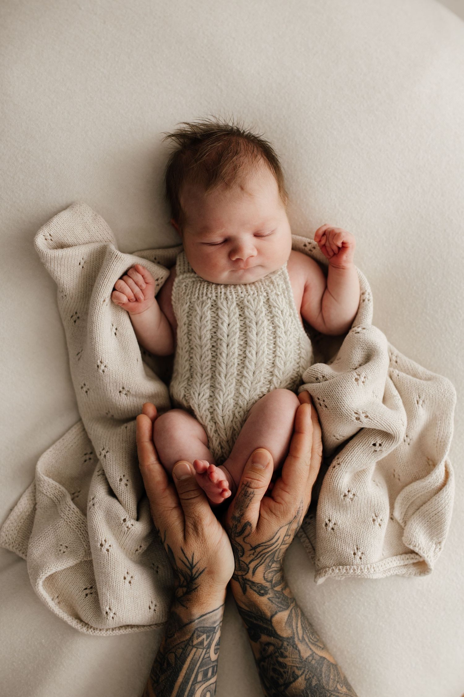 Newborn baby in knit romper lying on textured blanket with tattooed hands supporting.