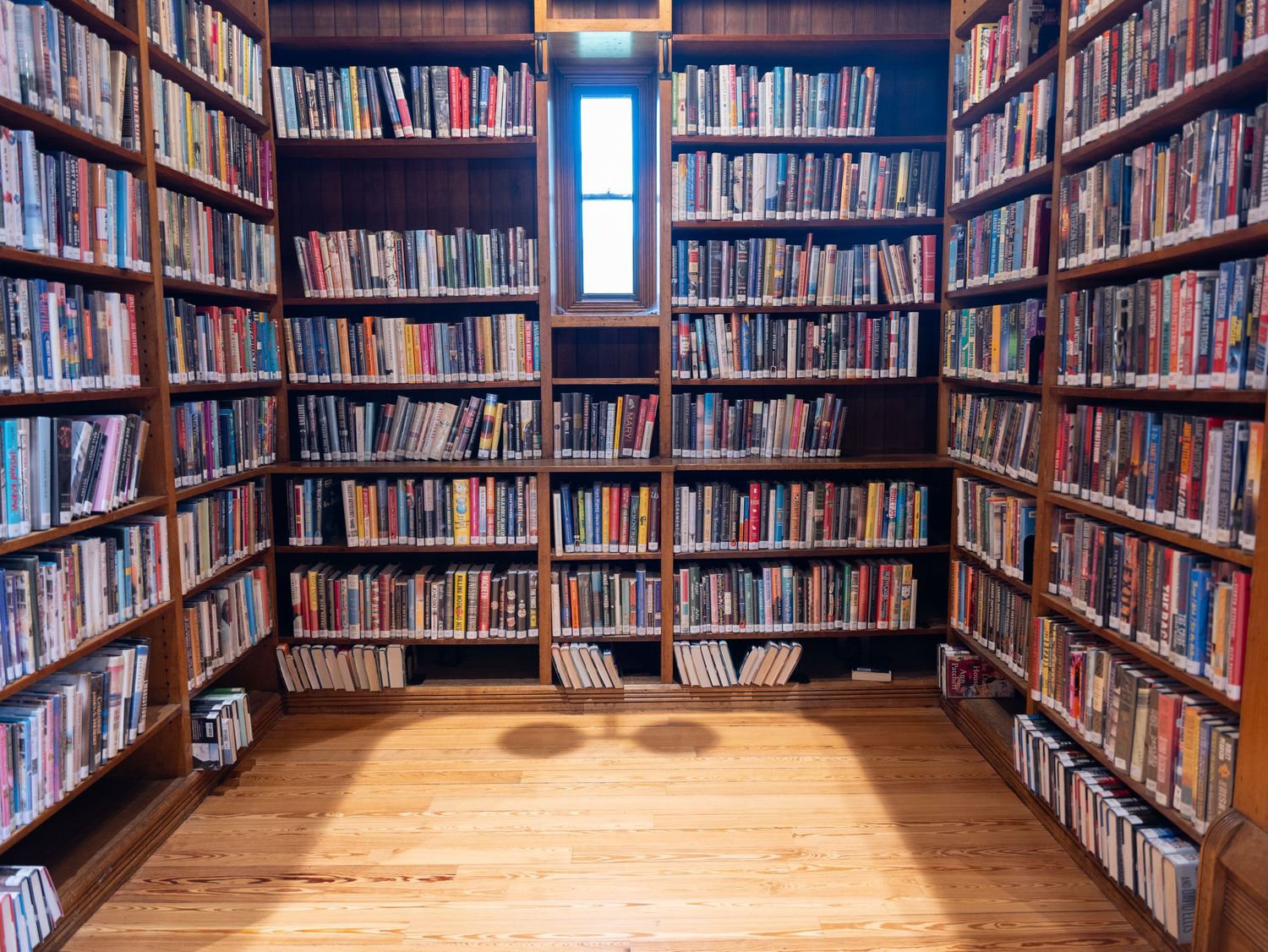 Floor-to-ceiling library shelves line the walls of a small room with wooden flooring and natural light.