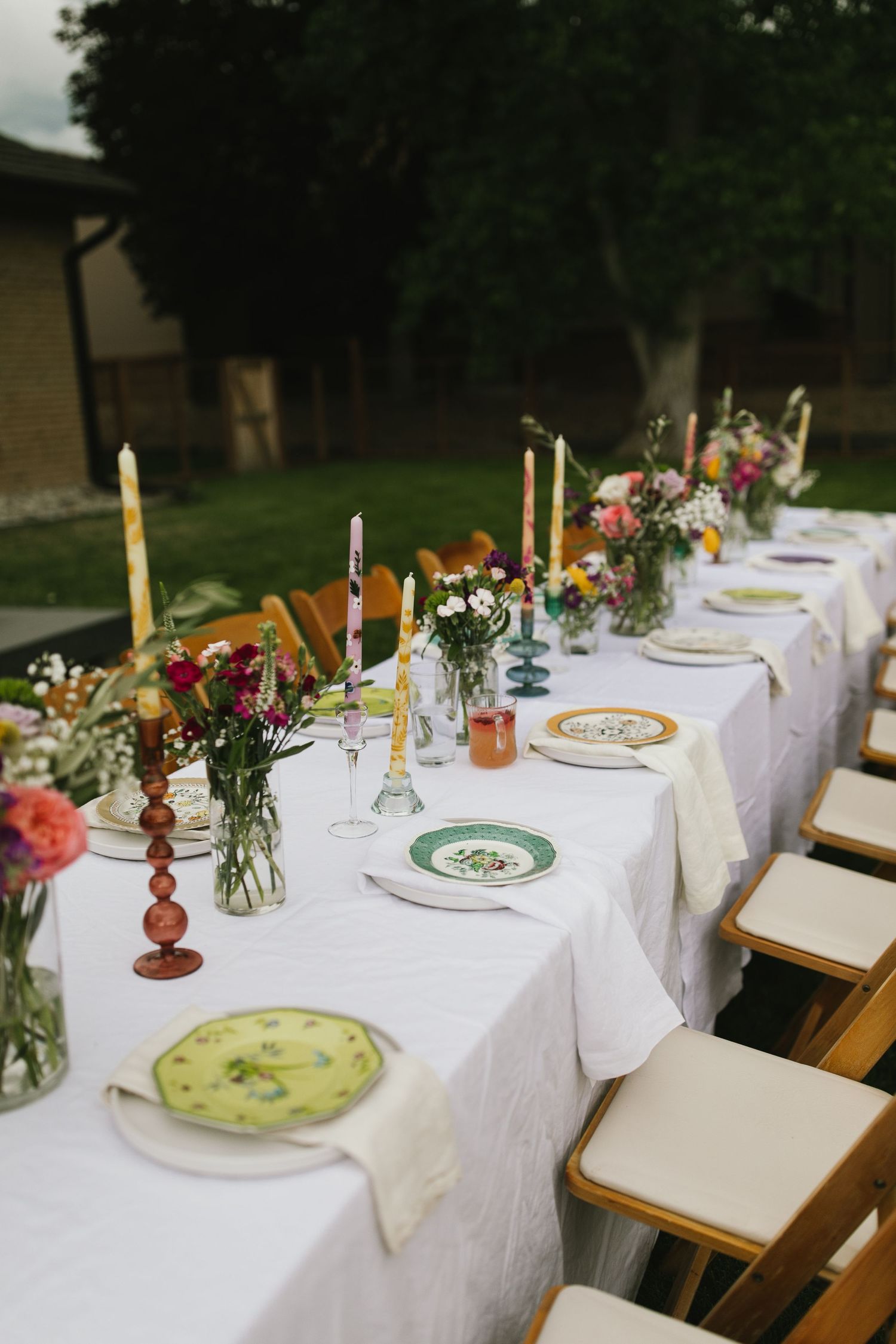 Elegant outdoor table setting with flowers, candles and colorful plates on white linen.