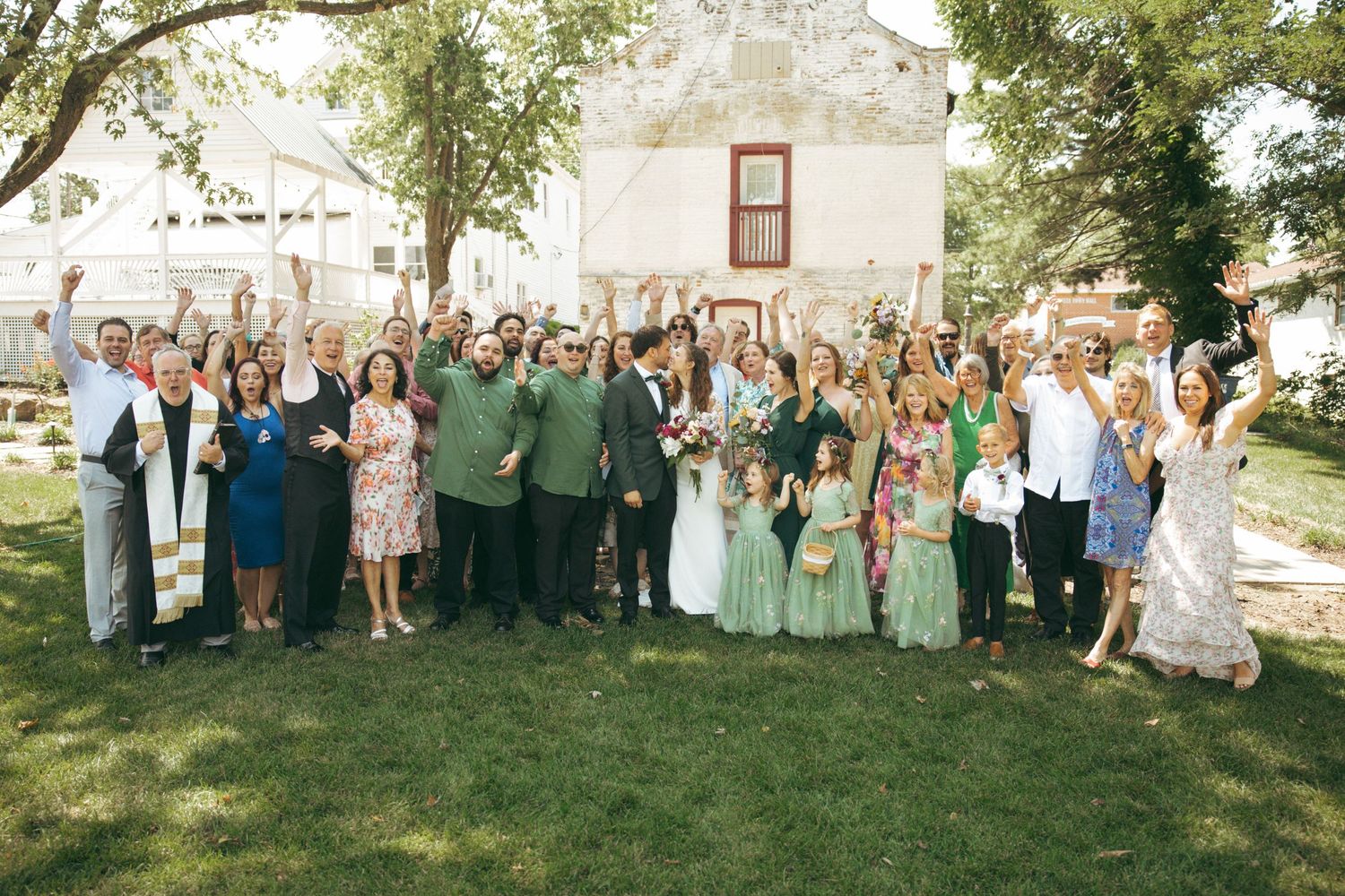 A large group celebrating at an outdoor wedding venue raises their arms in joy.