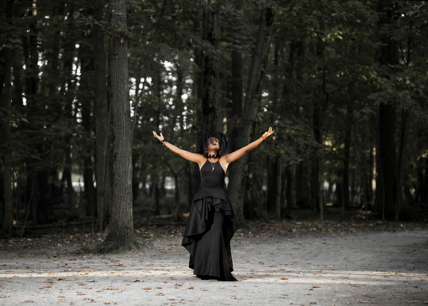 Figure in black dress with arms raised stands triumphantly in forest setting.
