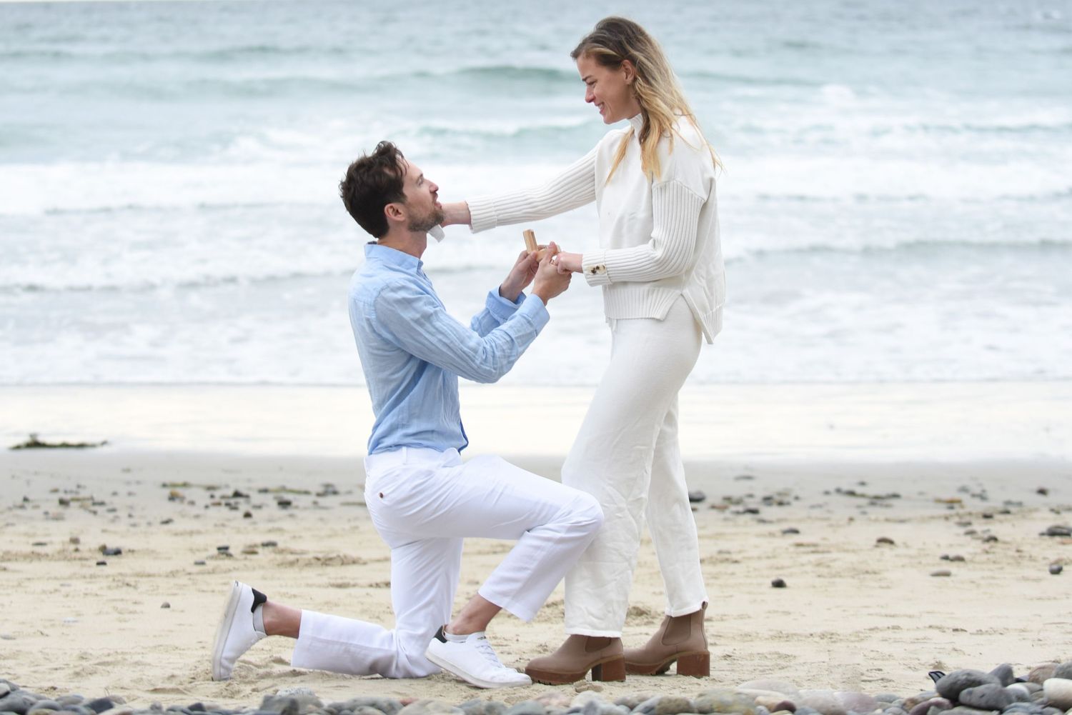 A marriage proposal moment captured on a scenic beach with coastal cliffs in the background.