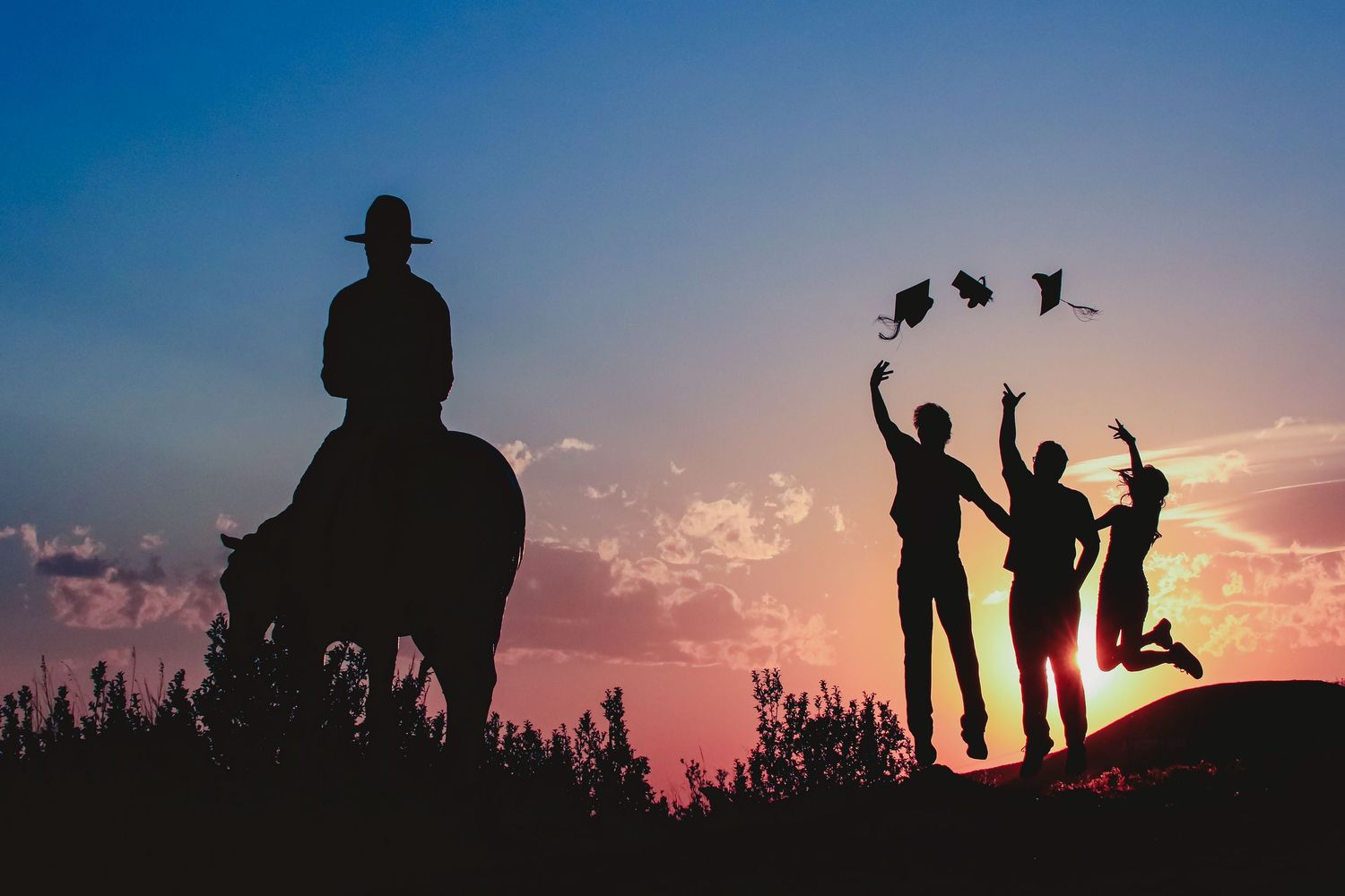 Silhouette of three graduates jumping and tossing caps at colorful sunset with equestrian nearby.