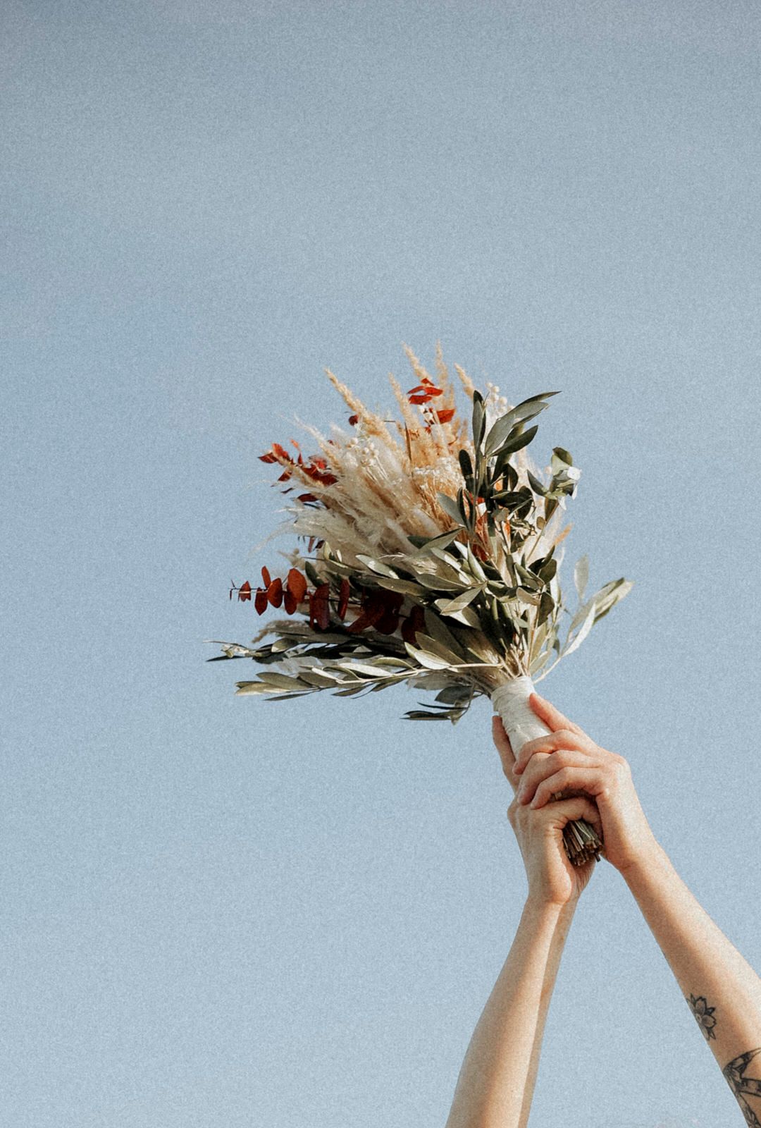 Hands raised against a blue sky holding a dried floral arrangement with white and brown flowers and green foliage.
