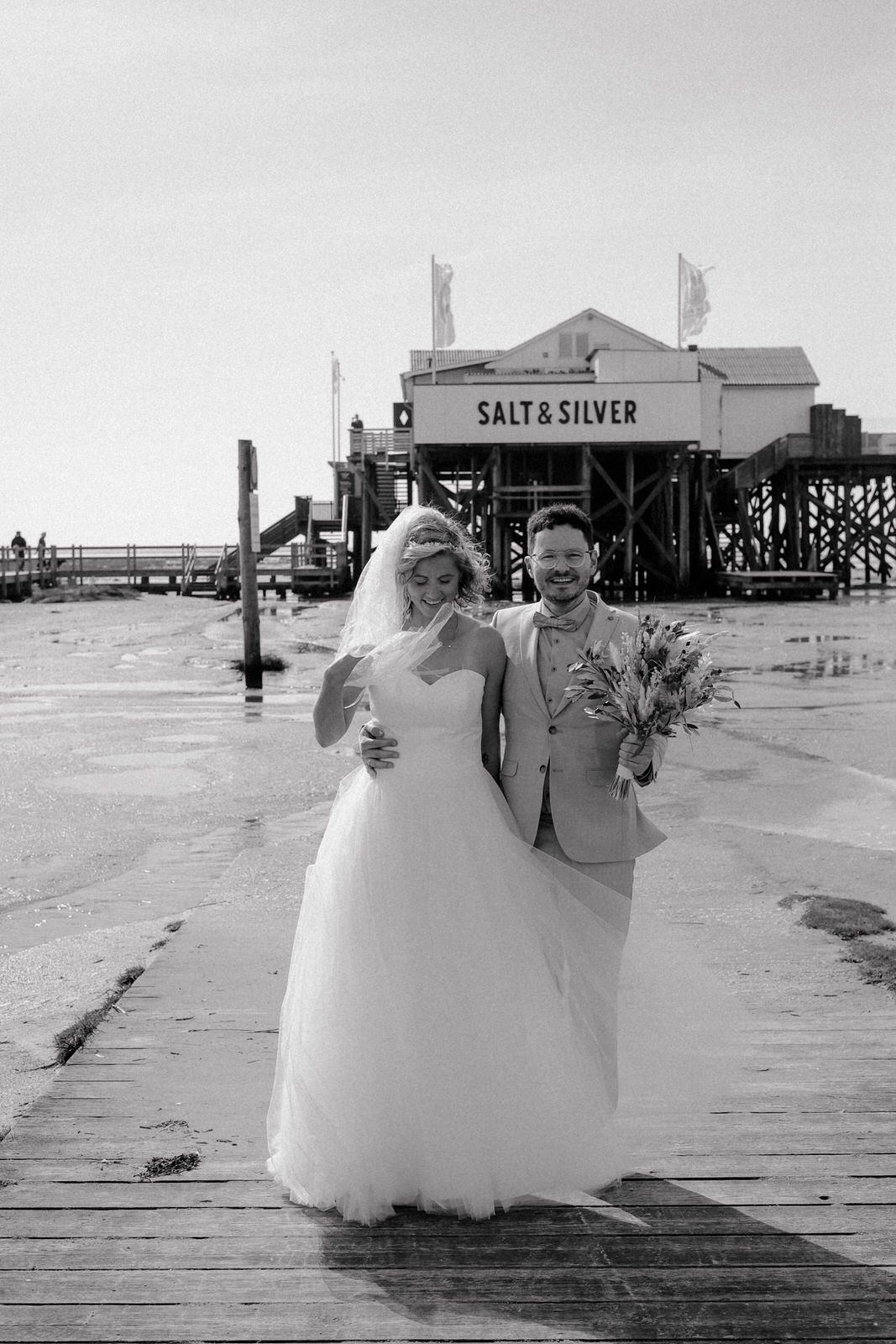 A couple walks together along a wooden pier at a beach with a vintage-style building in the background. Salt & Silver SPO.