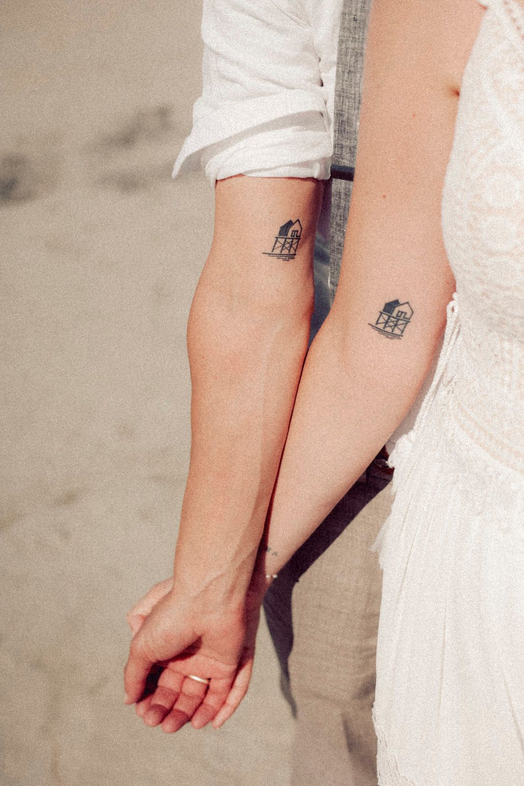 Close-up of two arms with matching small birdcage tattoos, wearing white clothing against a neutral background.