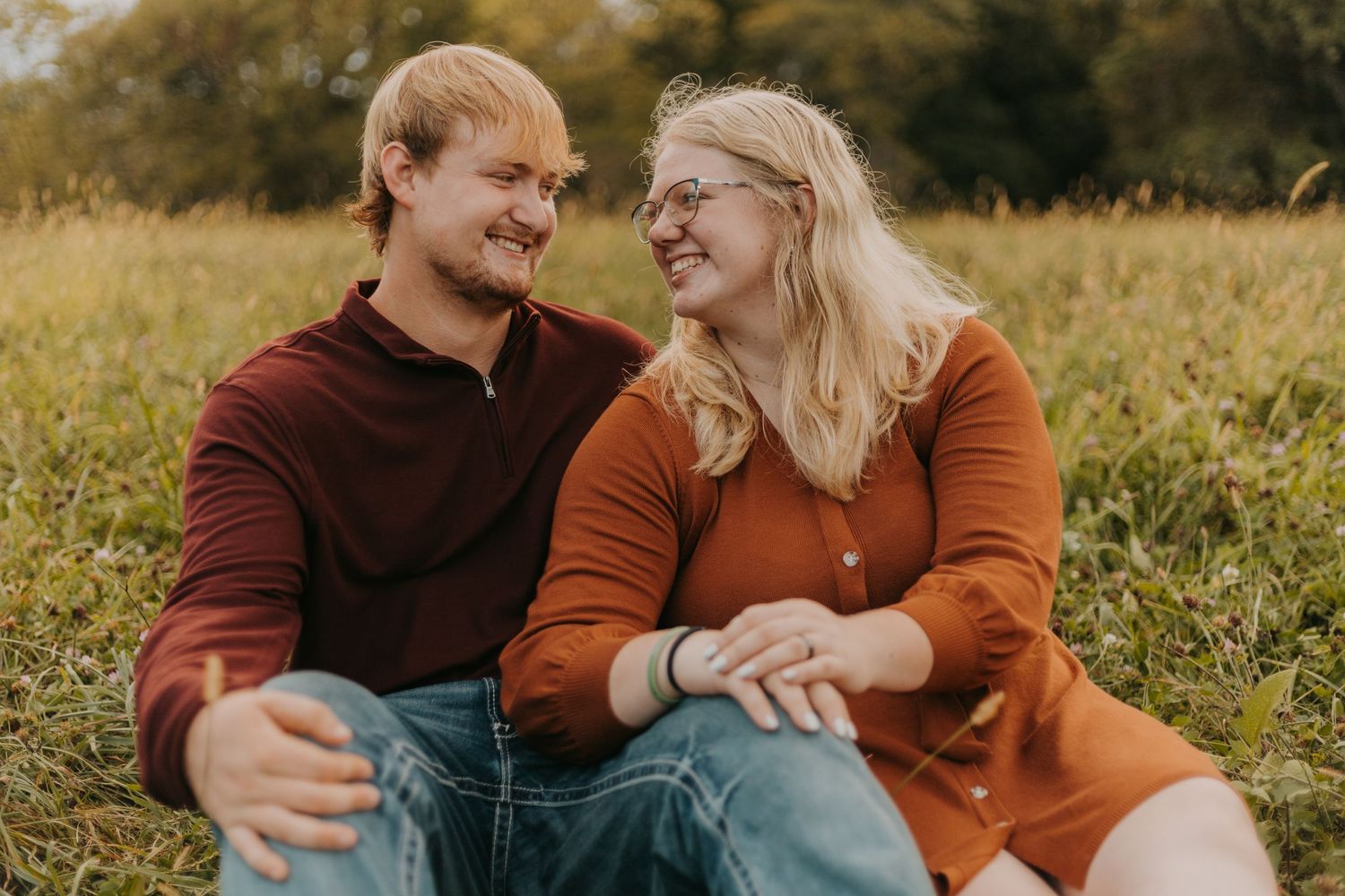 A couple sits together laughing in a sunlit autumn field wearing coordinating burgundy and orange sweaters.