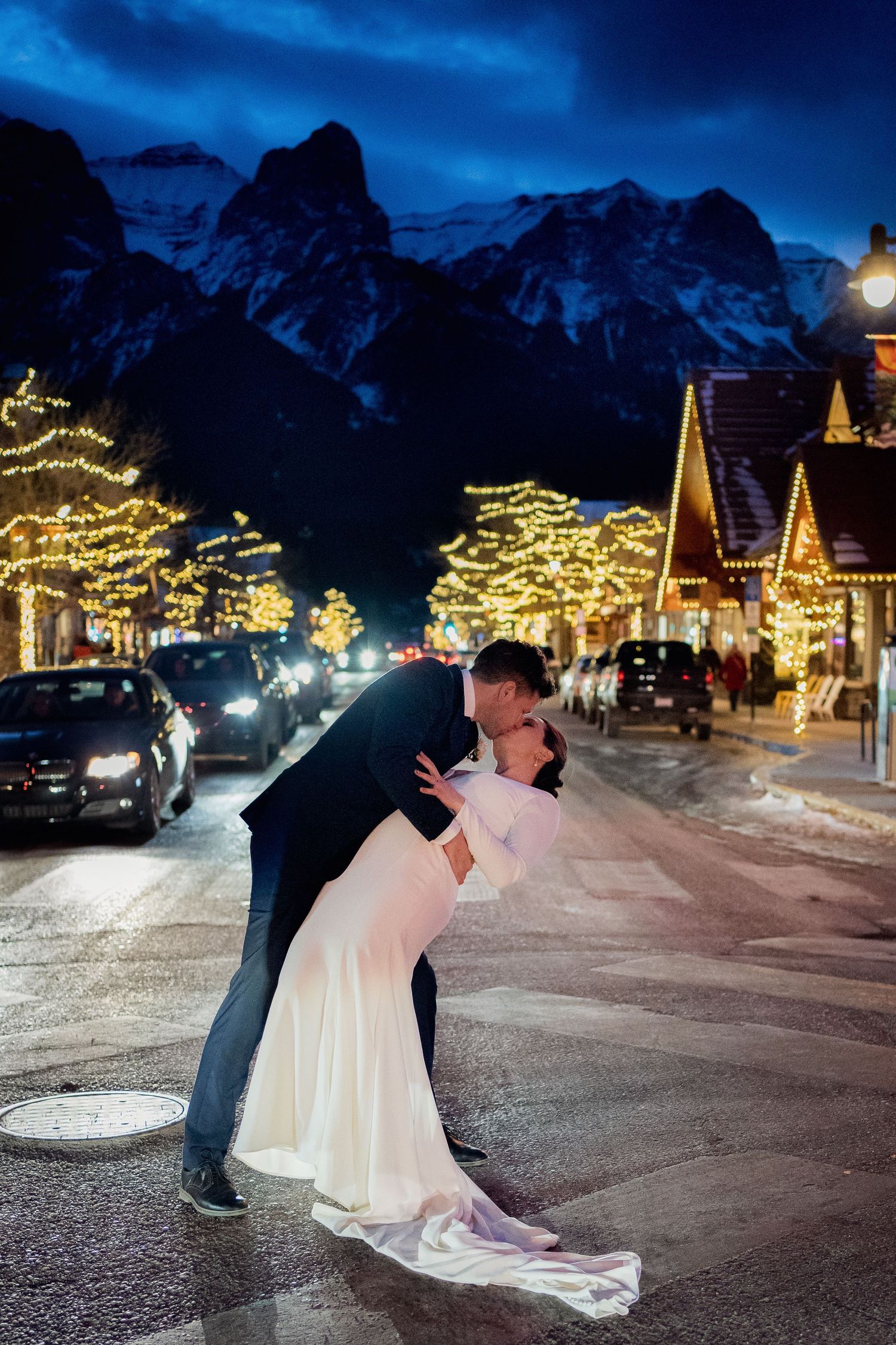 A romantic winter wedding photo with mountain backdrop and festive holiday lights in a snowy village.