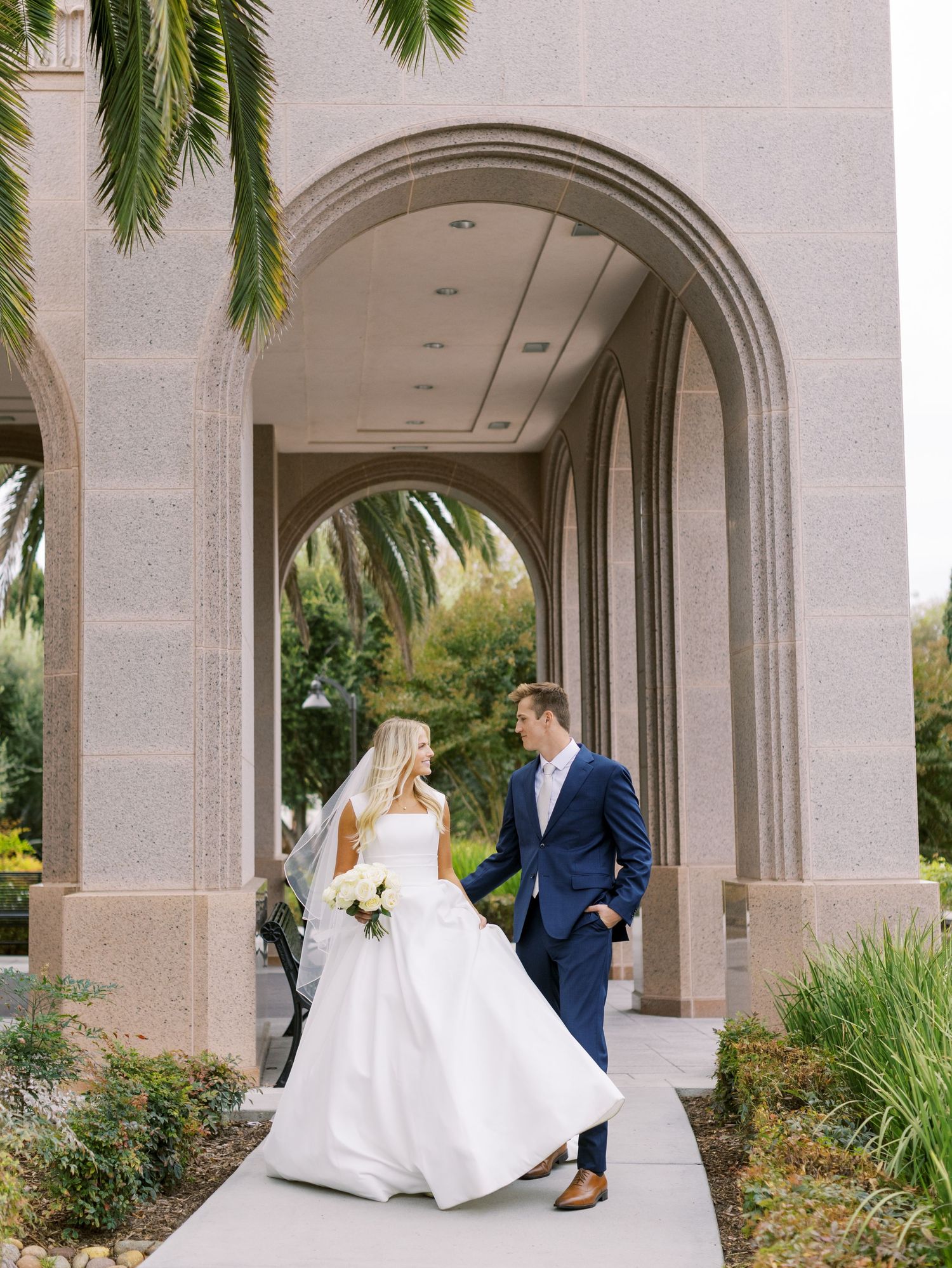 Elegant couple stands in archway with palm trees and landscaping at grand stone building entrance.