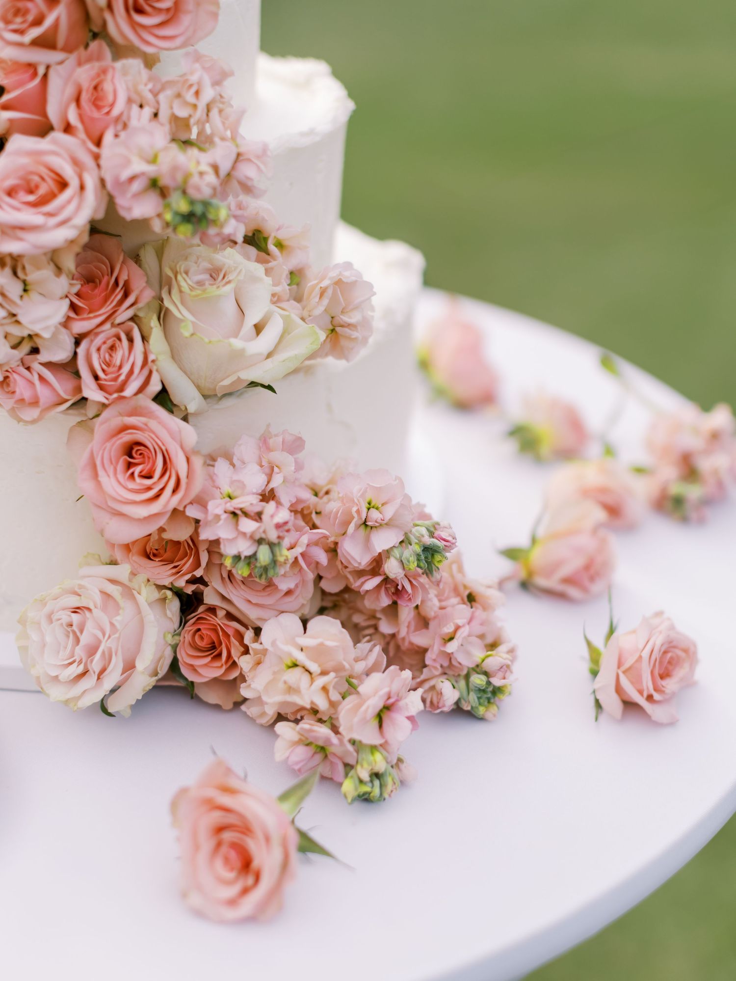 White tiered wedding cake decorated with cascading pink roses and hydrangeas against a blurred green background.