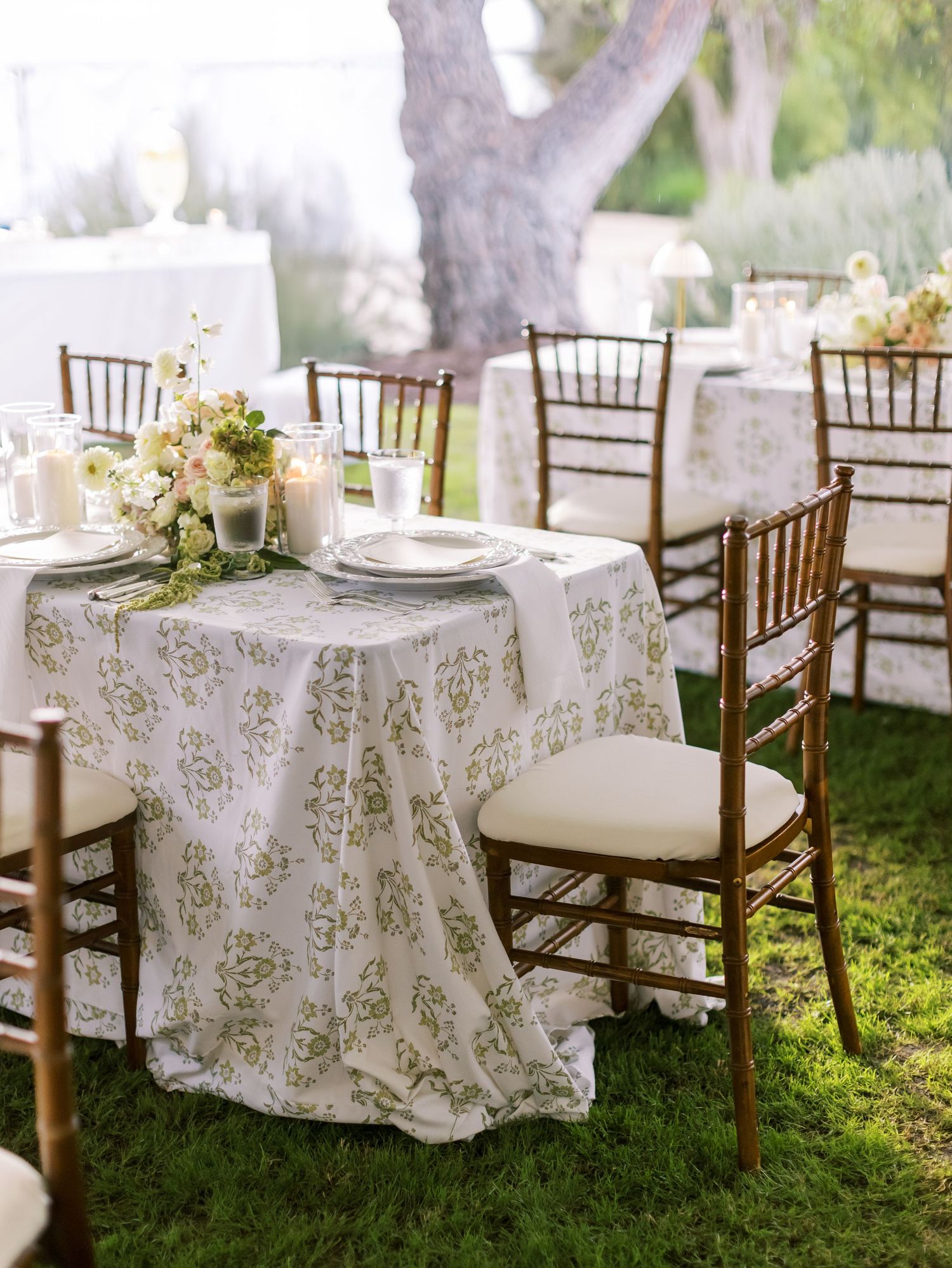 Elegant outdoor wedding table setting with floral patterned tablecloth and dark wood Chiavari chairs on green grass.