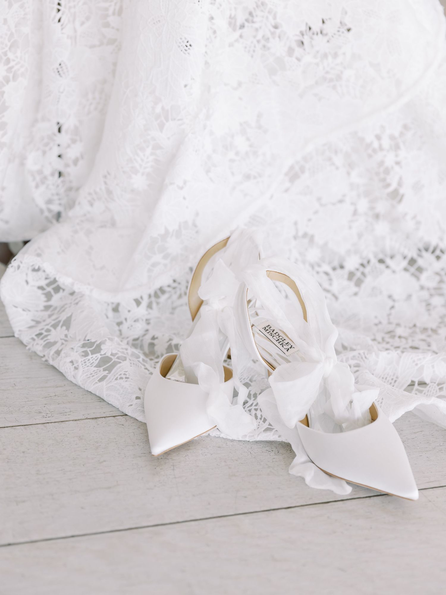 Elegant white bridal shoes with pointed toes sit beside delicate lace fabric on a light wooden floor.