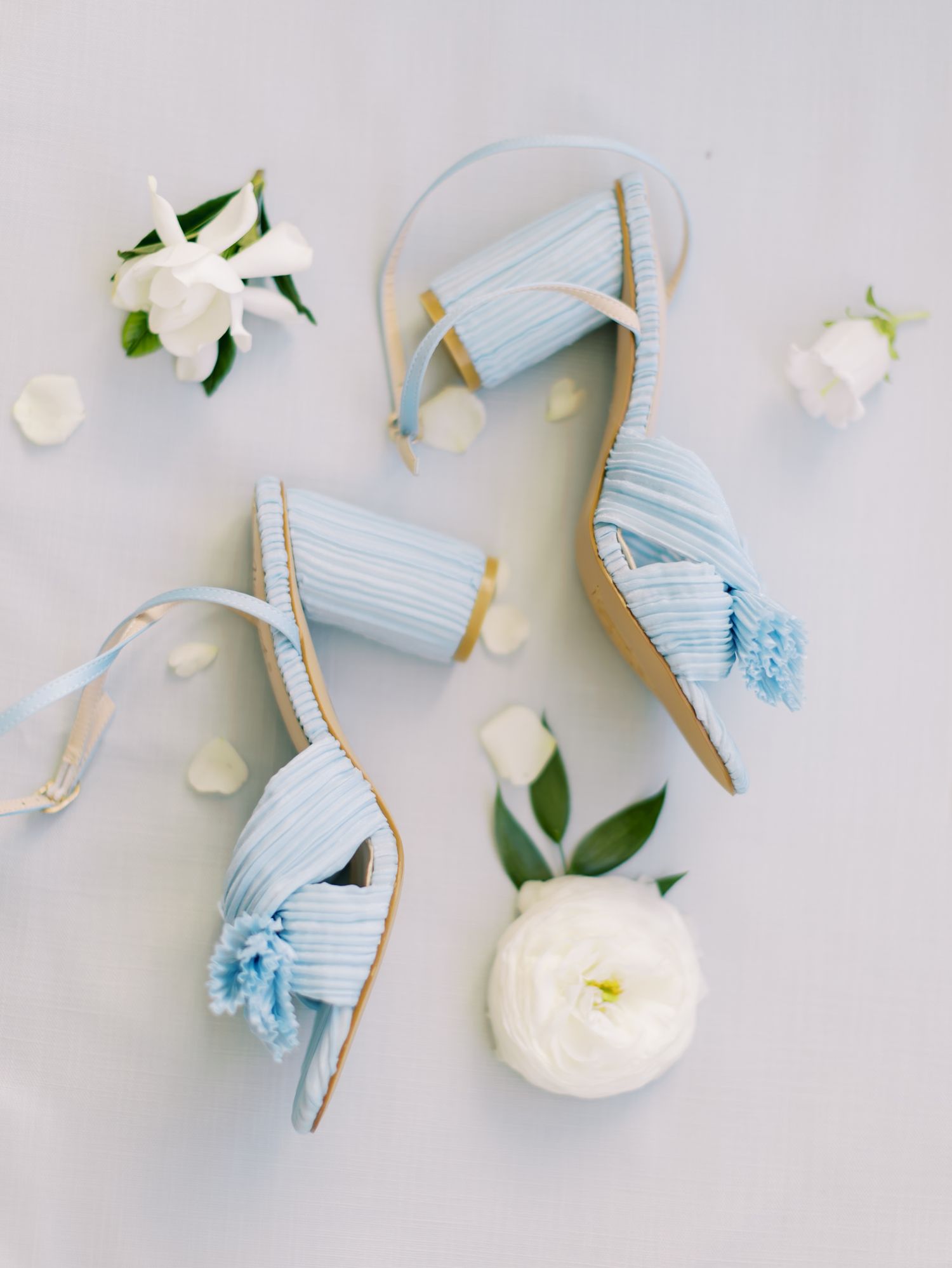 Light blue wedding sandals with delicate straps styled with white garden roses and petals on a soft gray background.