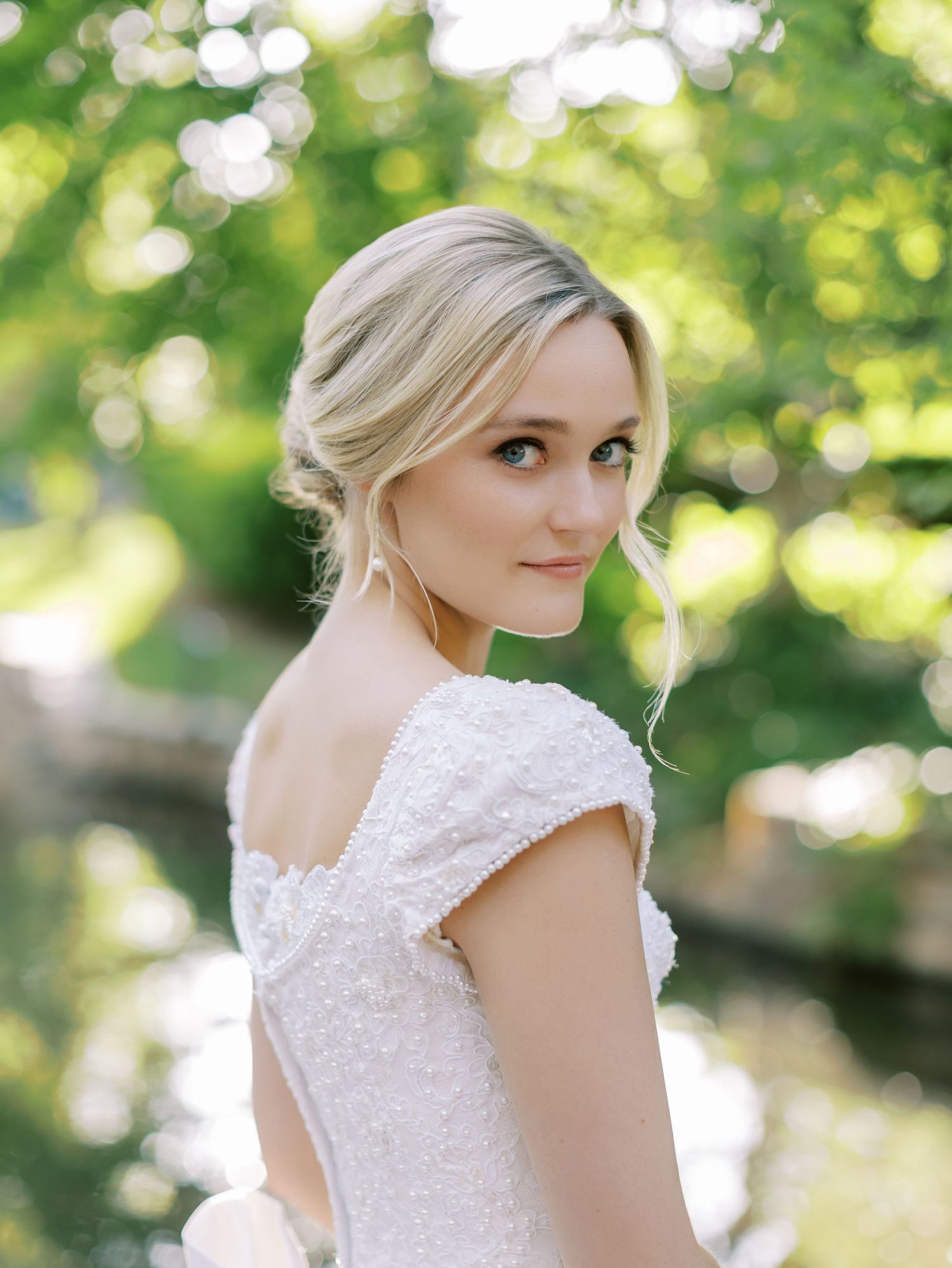 Bride in white lace cap sleeve wedding dress poses outdoors with soft green bokeh background in natural light.