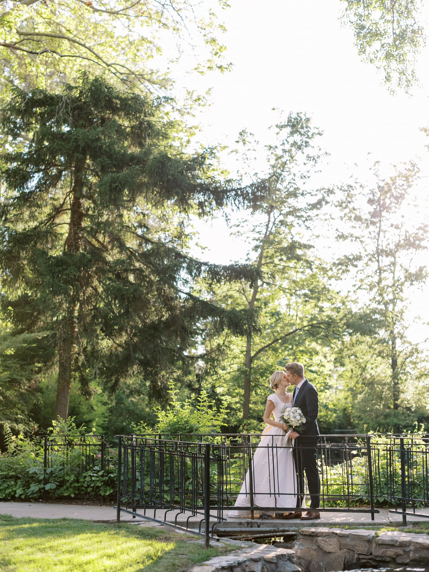 A couple stands romantically on a garden bridge surrounded by lush evergreen trees on a sunny summer day.