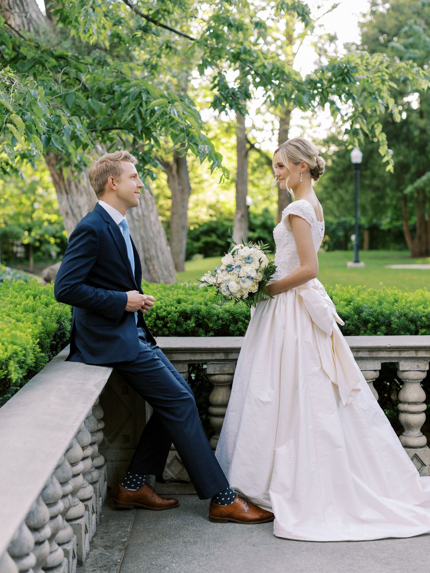 A couple in wedding attire stands together on a stone balcony in a lush garden setting during their special day.