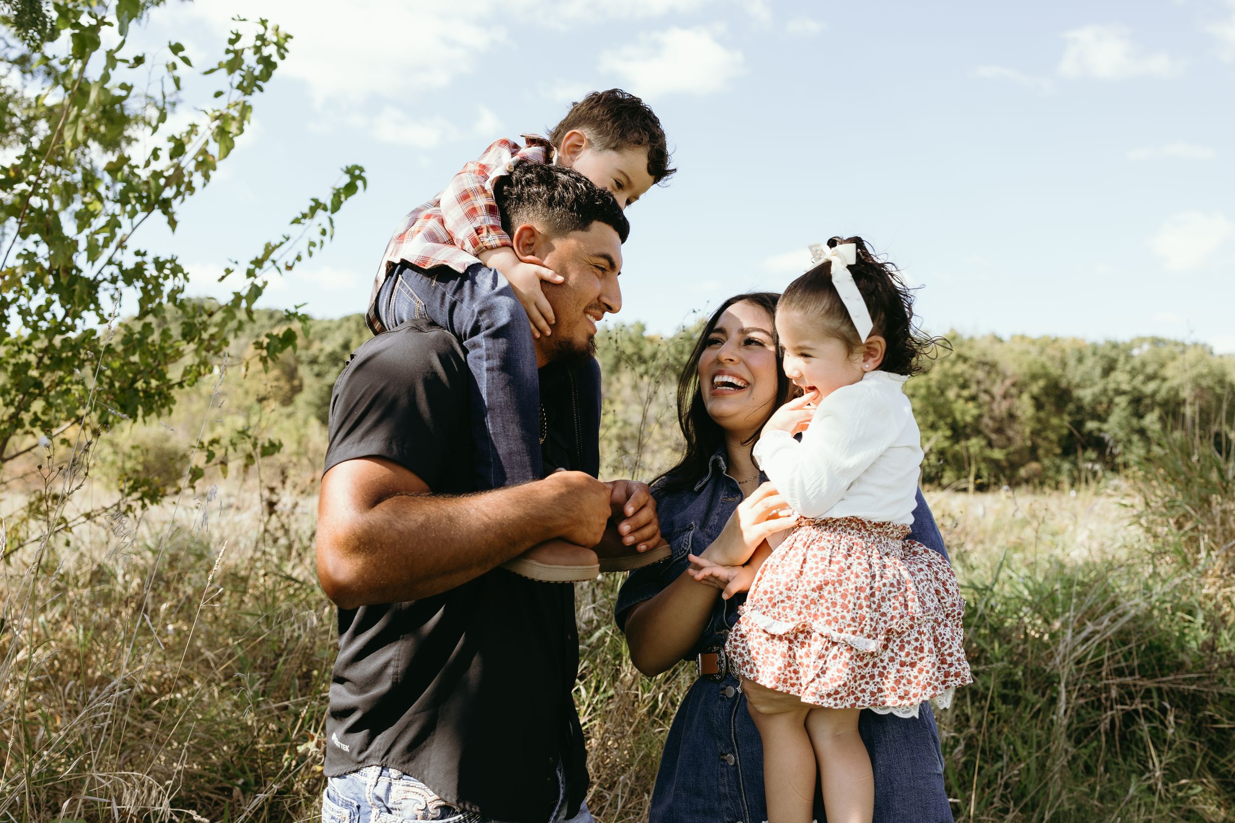 A joyful family embraces and laughs together while enjoying a sunny day in a natural outdoor setting.