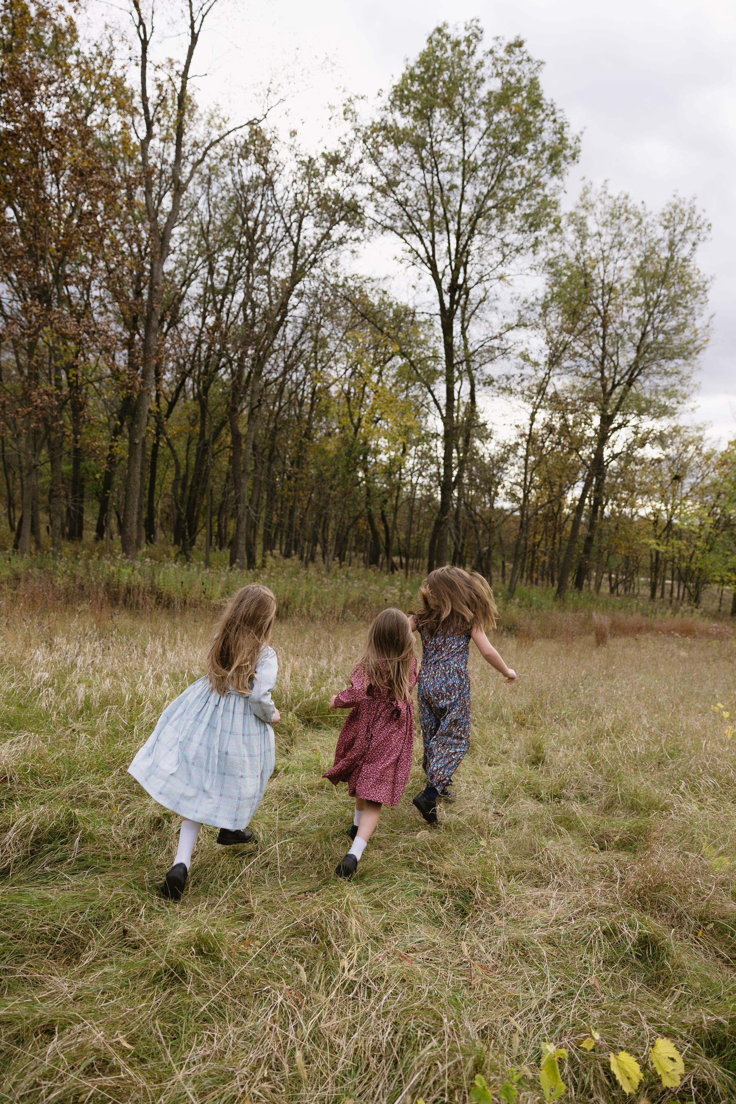 Three children in dresses running through a grassy field on an autumn day with tall trees in the background.