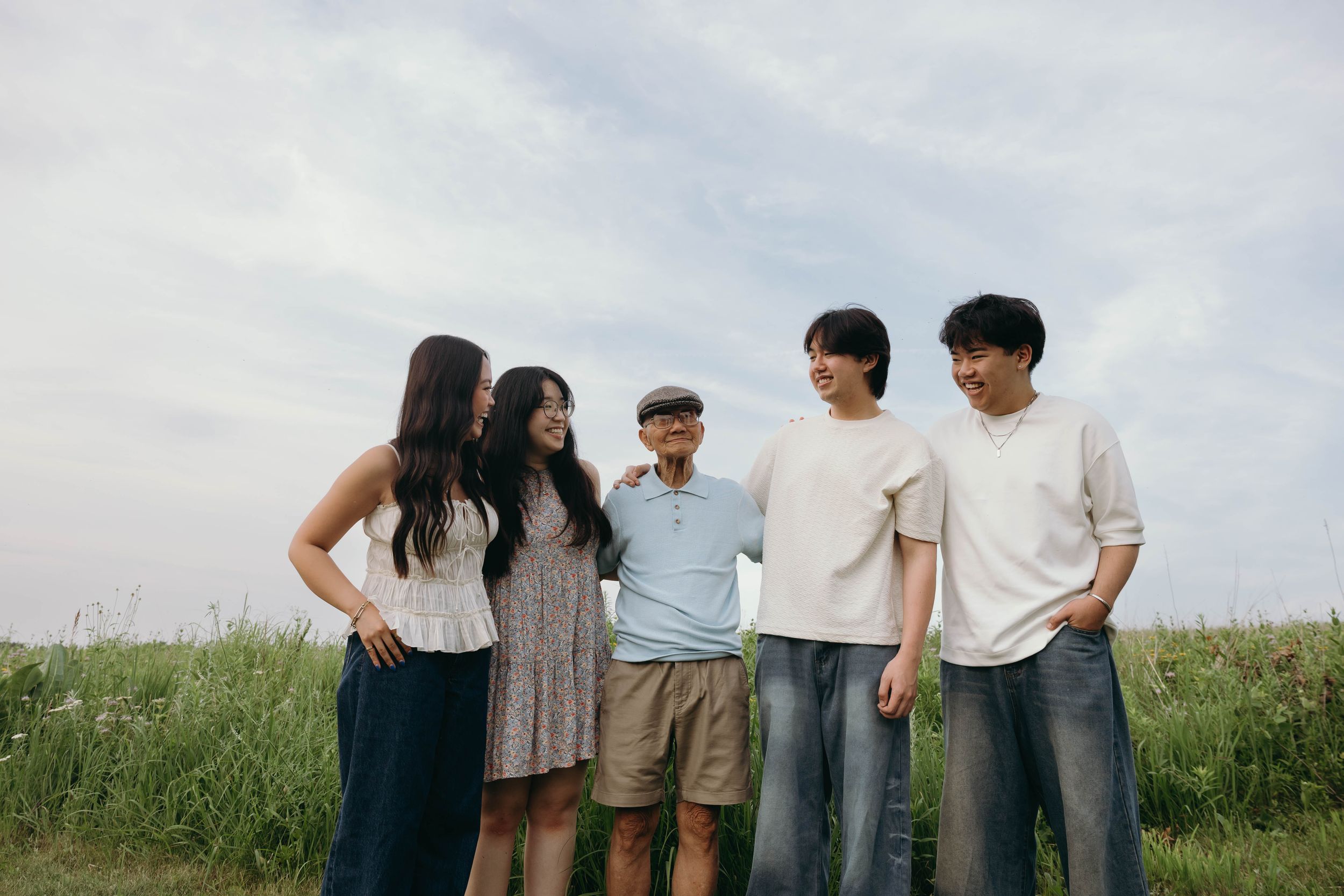 Group of friends in casual clothing standing together in a grassy field against a cloudy sky backdrop.