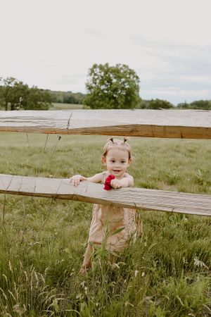 Small child in pink outfit peeks through wooden fence rails in grassy field.
