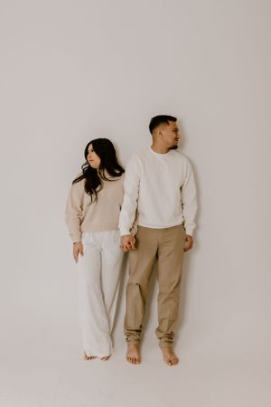 A minimalist portrait of a couple in matching cream sweaters holding hands against a neutral wall.