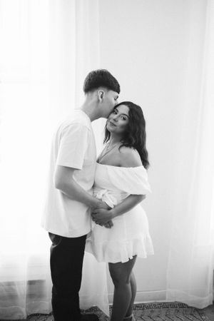 Intimate black and white portrait of a couple in white clothing embracing against a bright wall.