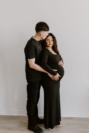 Intimate maternity portrait of couple in black attire against white backdrop.