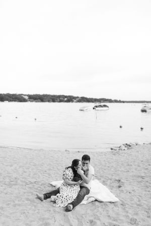 A couple sitting together on a sandy beach near the water in black and white.