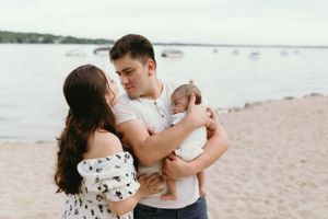 A couple embraces while holding their baby on a beach with boats in the background.