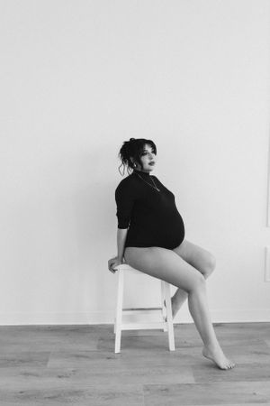 Artistic maternity photo featuring expecting mother in black sweater posed on white stool.