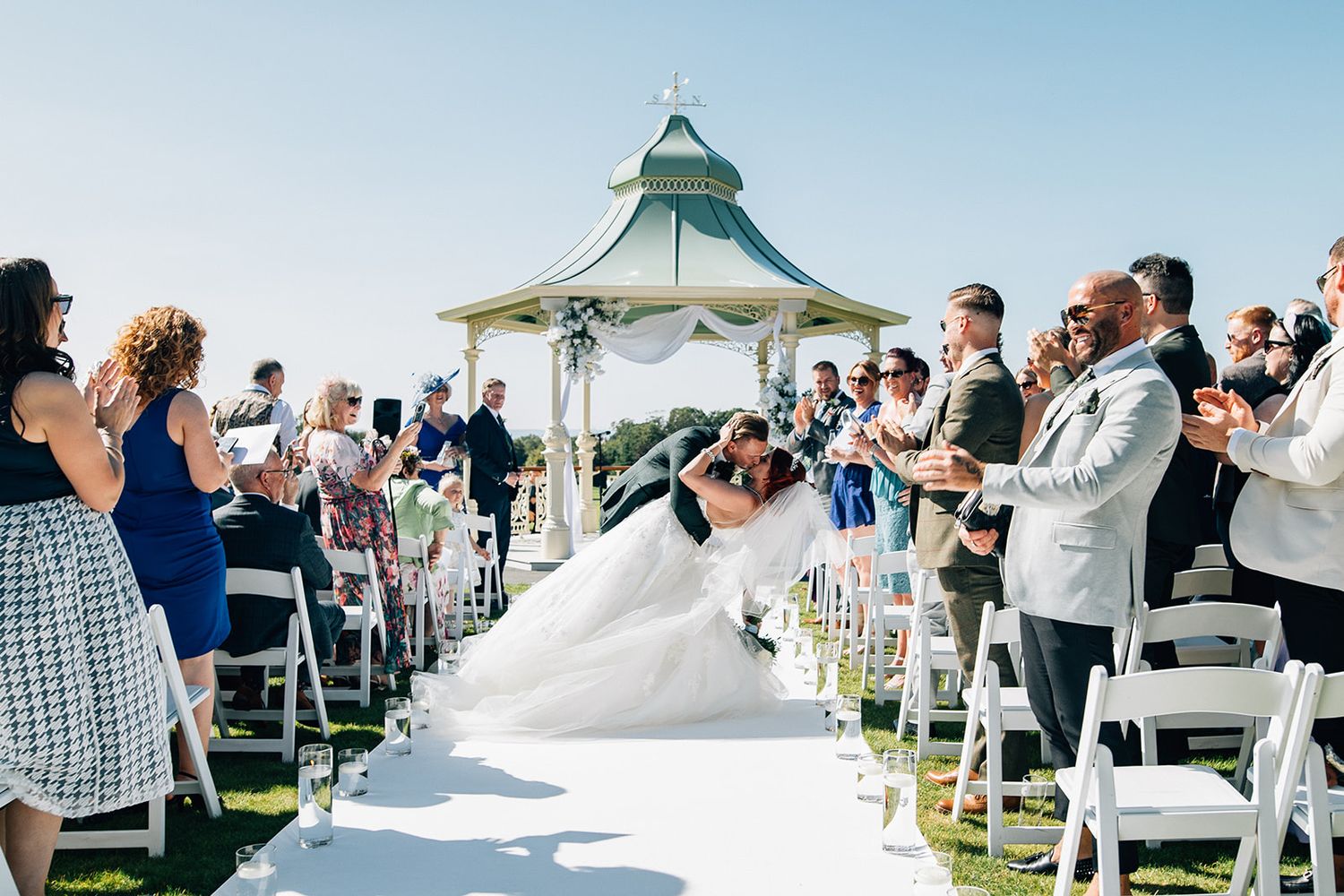 Wedding ceremony taking place under a gazebo with guests seated on white chairs outdoors.