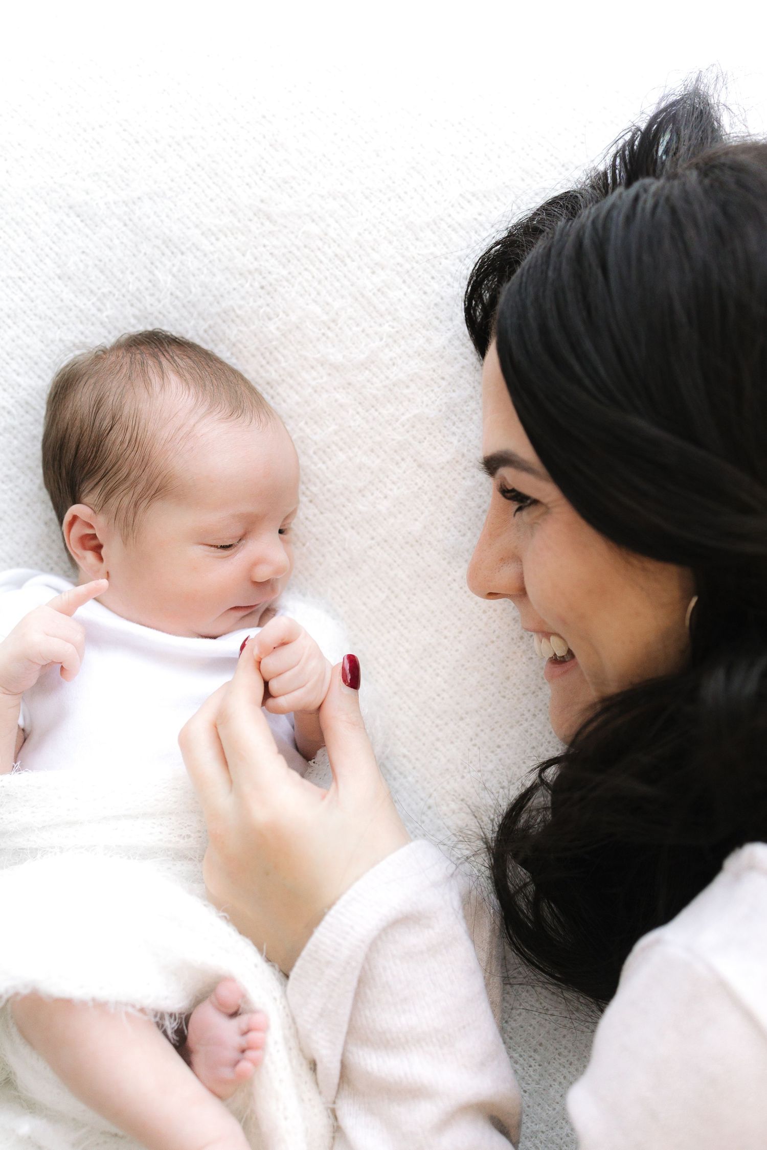 A smiling dark-haired mother lying down, lovingly gazing at her newborn baby in a white outfit.