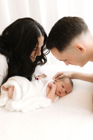 New parents lovingly gaze at their newborn baby wrapped in white on a soft white blanket.