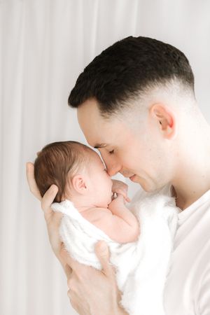 A father tenderly holds his newborn baby wrapped in a white blanket, touching foreheads in a gentle moment.