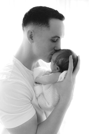 Father tenderly kisses his newborn baby wrapped in a white blanket in black and white photo.