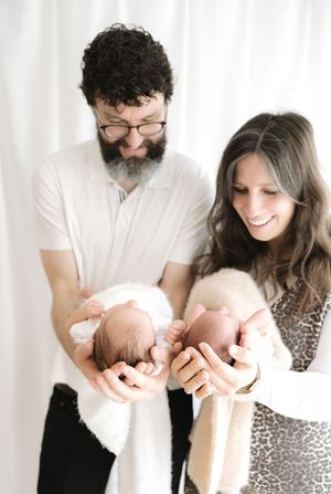 Parents smiling and cradling two newborn twins in their hands against a white backdrop.